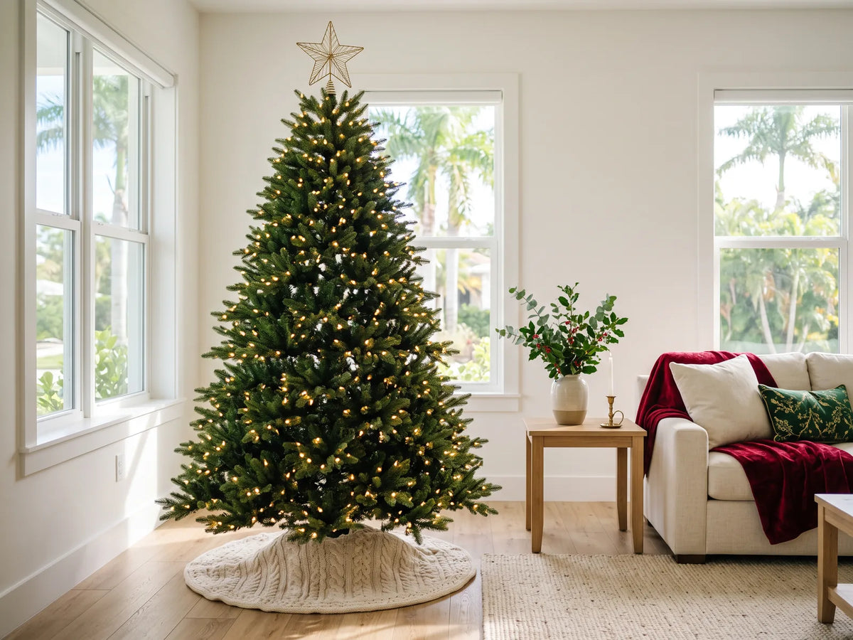 Tall artificial Christmas tree with warm white lights, gold star and cream knit skirt by windows with palms